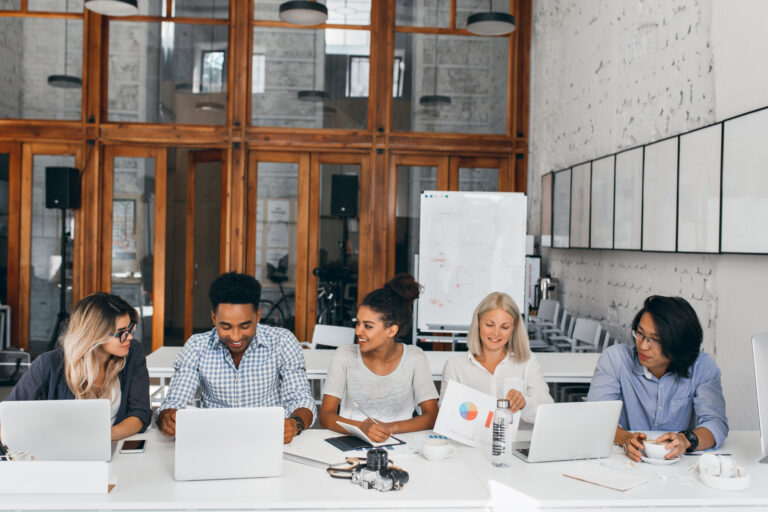 Tired asian it-specialist drinking coffee and watching female colleague working with laptop. Indoor portrait of young business people sitting at the table together in conference hall.
